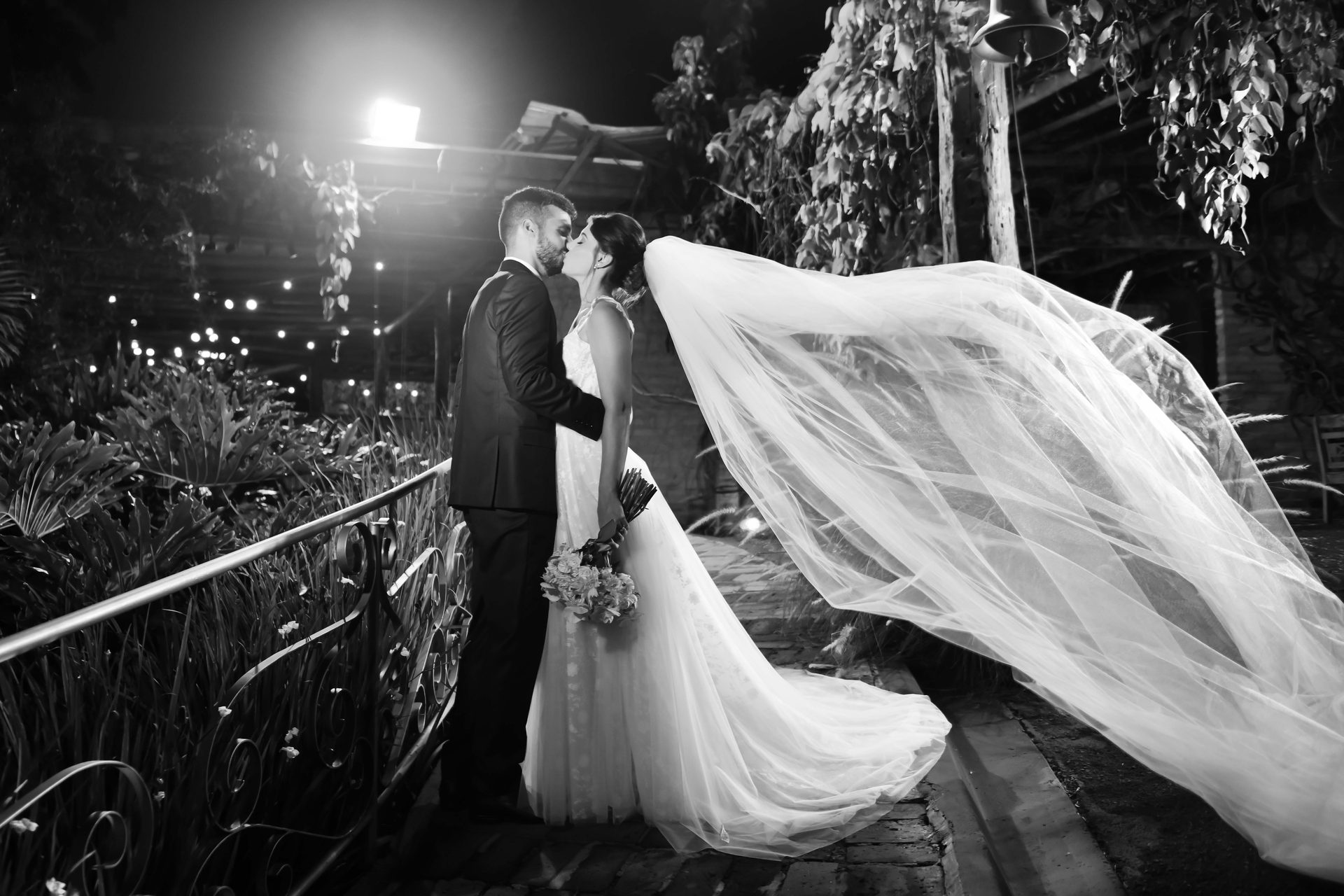 Fotografia em preto e branco de casal de noivos com o véu ao vento à noite